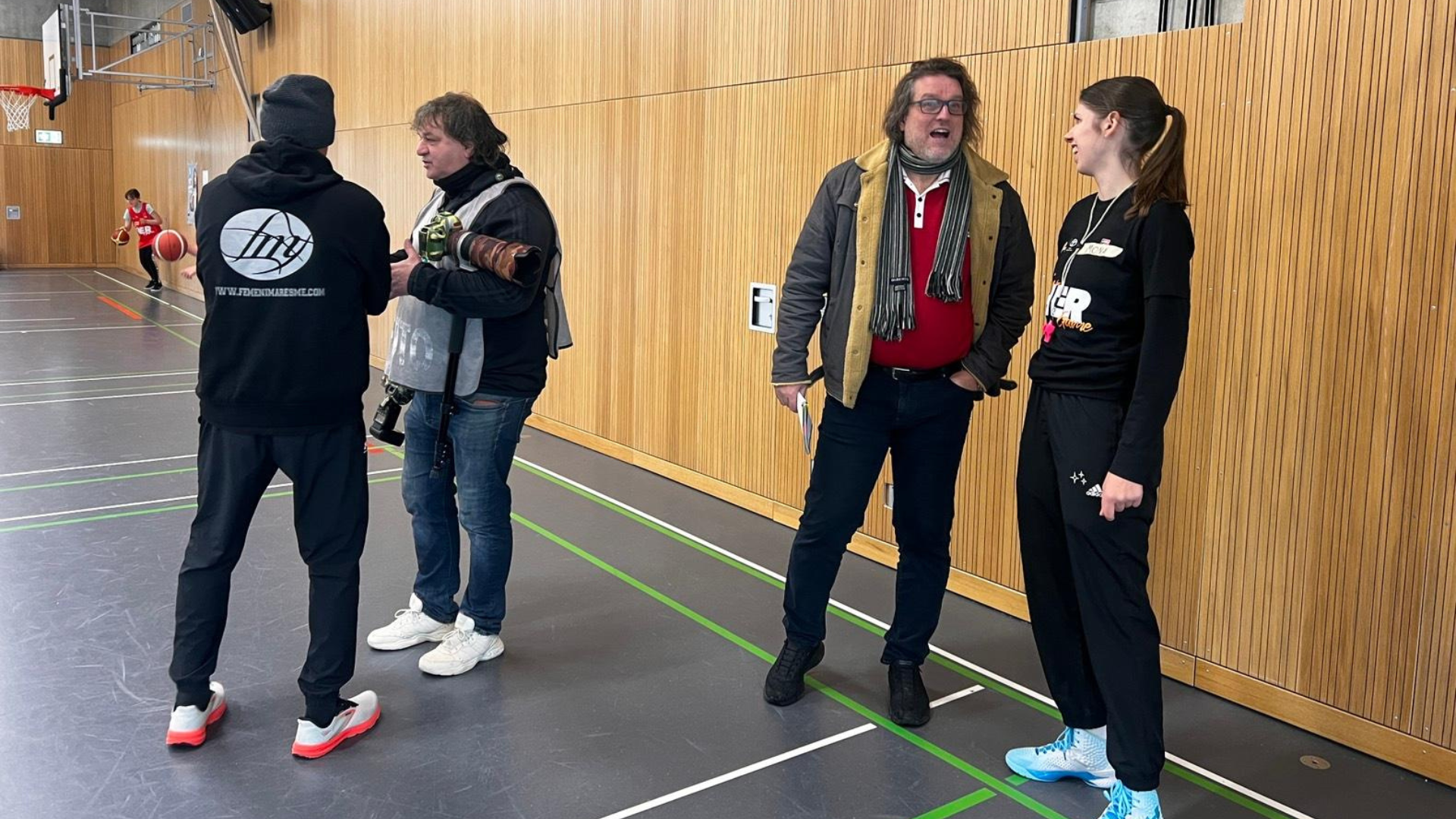 Fotograf, Besucher und Trainer unterhalten sich in einer Sporthalle während Mädchen im Hintergrund Basketball spielen