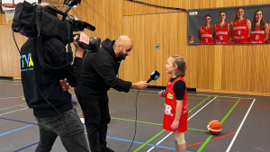 Mädchen wird mit Mikrofon und Kamera in einer Basketballhalle interviewt